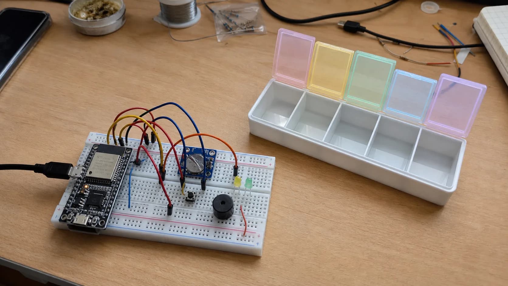 Prototype smart medicine reminder box with an ESP32, RTC module, buzzer, LEDs, and a multi-slot pill box on a workbench.