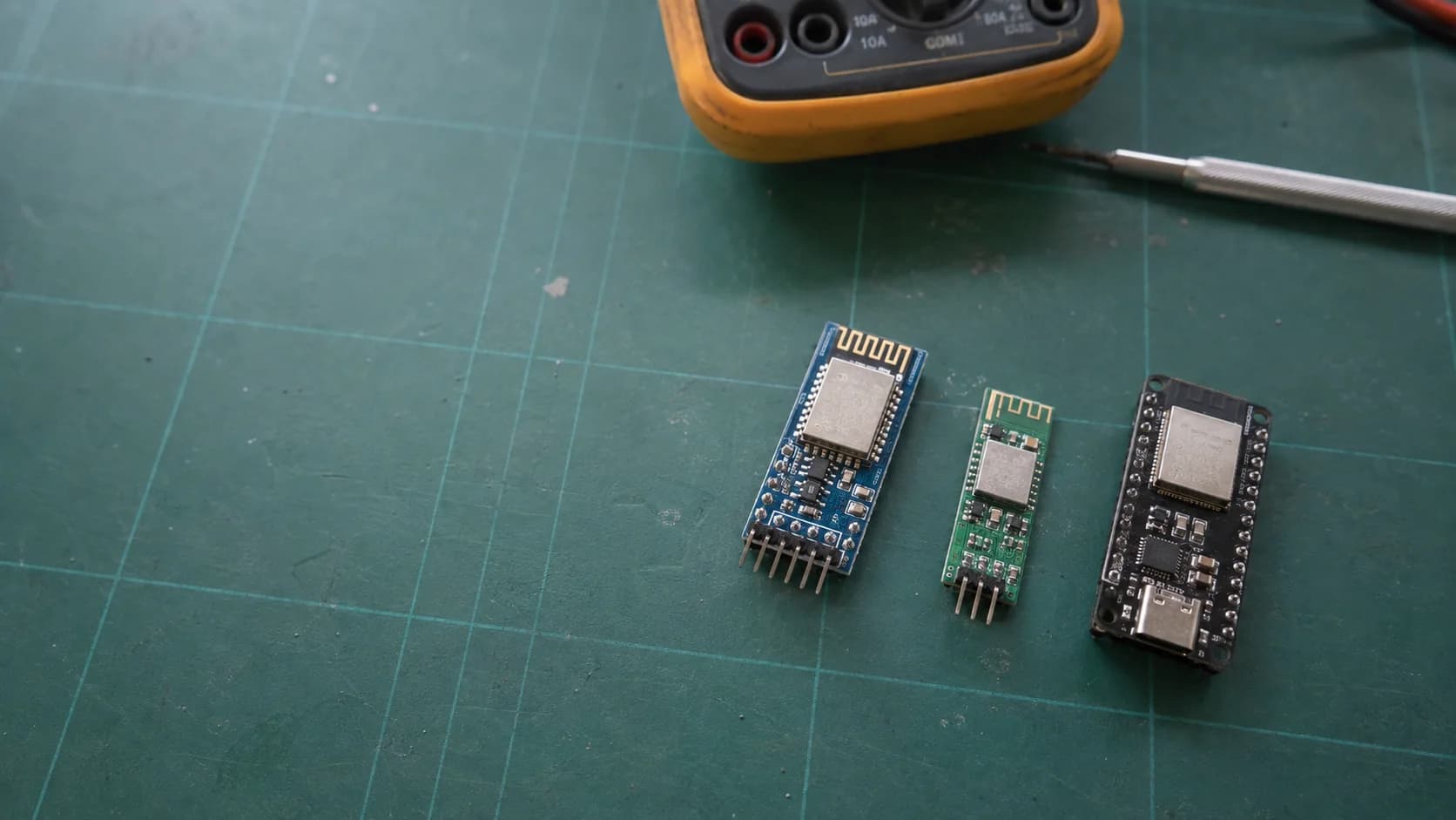 Three Bluetooth module boards arranged on a used ESD workbench beside a multimeter and precision screwdriver