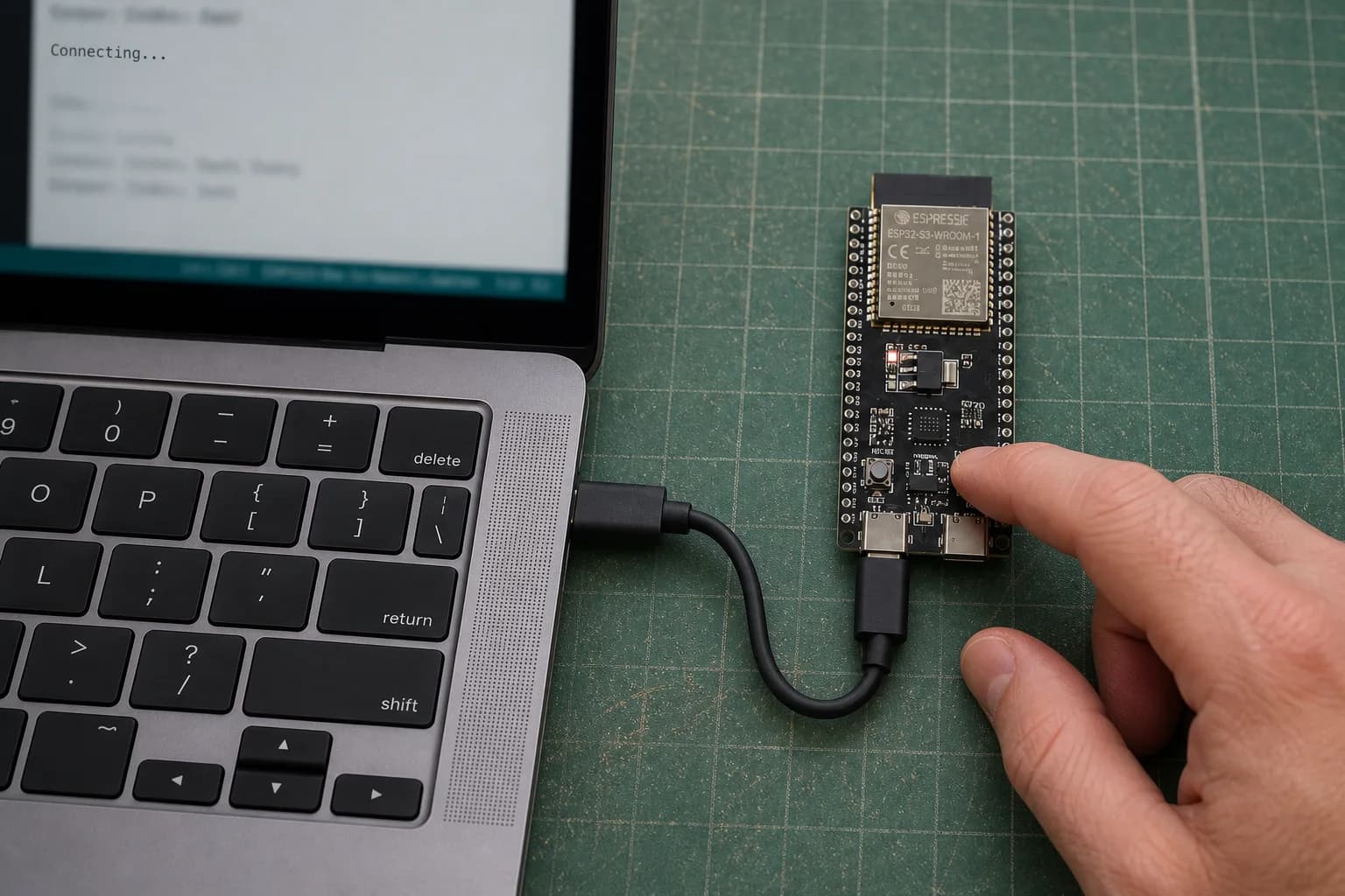 A hand holds the BOOT button on an ESP32-S3-DevKitC-1 while its USB cable remains connected and a MacBook Pro waits for the device.