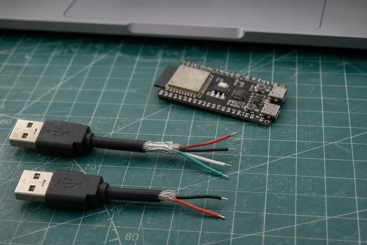 Two stripped USB cable samples on an electronics bench: a four-wire data cable and a two-wire charge-only cable beside an ESP32-S3 DevKitC and MacBook Pro.