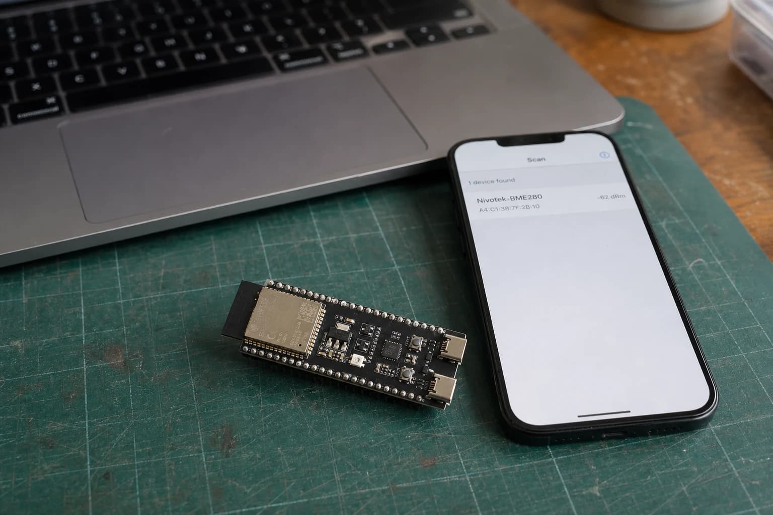 An ESP32-S3 development board and smartphone testing Bluetooth Low Energy beside a MacBook Pro on a realistic electronics workbench