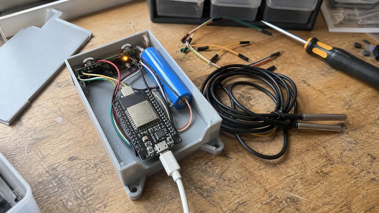 Prototype fridge temperature alert device with an ESP32 board, small backup battery, 3D-printed enclosure, and two waterproof DS18B20 probes on a workbench.