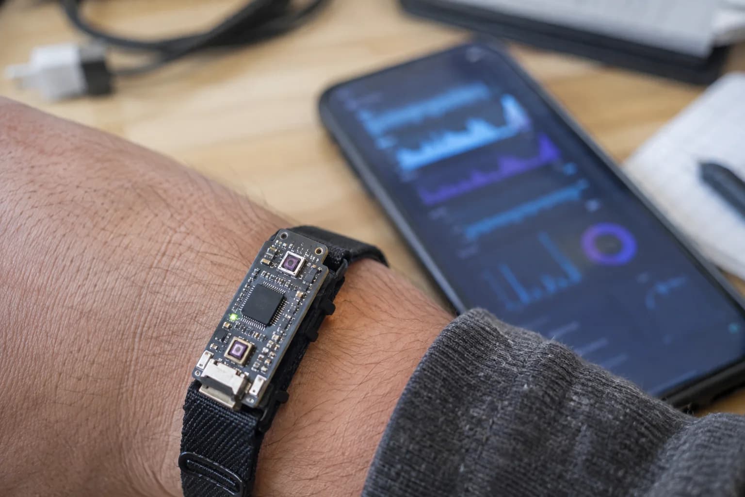 Small BLE wearable strapped to a wrist with a blurred phone showing sensor graphs in the background.
